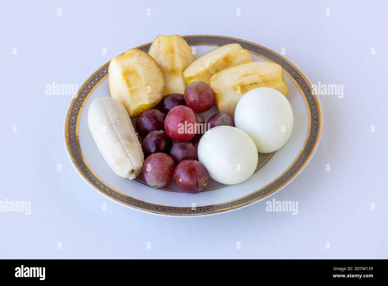 Eine Keramikplatte mit einer bunten und nahrhaften Auswahl an geschälten Apfelscheiben, roten Trauben, Banane und zwei hart gekochten Eiern; ein gesundes, High-Pro Stockfoto