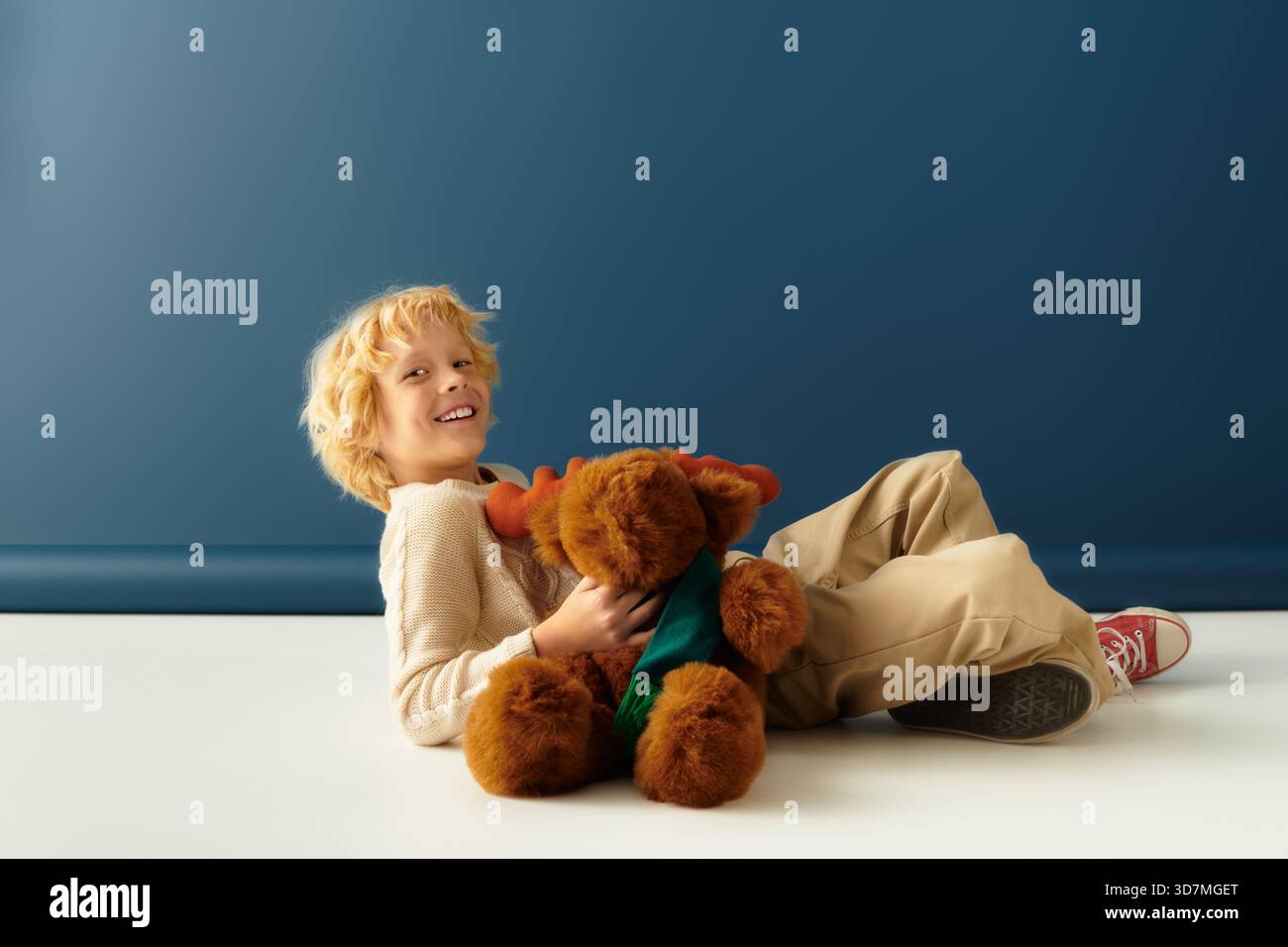 Ein blonder Junge lacht mit einem Plüschtier in einem Studio mit blauem Hintergrund. Stockfoto