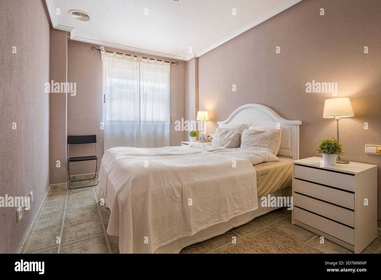 Gemütliches Schlafzimmer mit weißen Möbeln, sanfter Beleuchtung, transparenten Vorhängen und beruhigenden neutralen Wänden Stockfoto