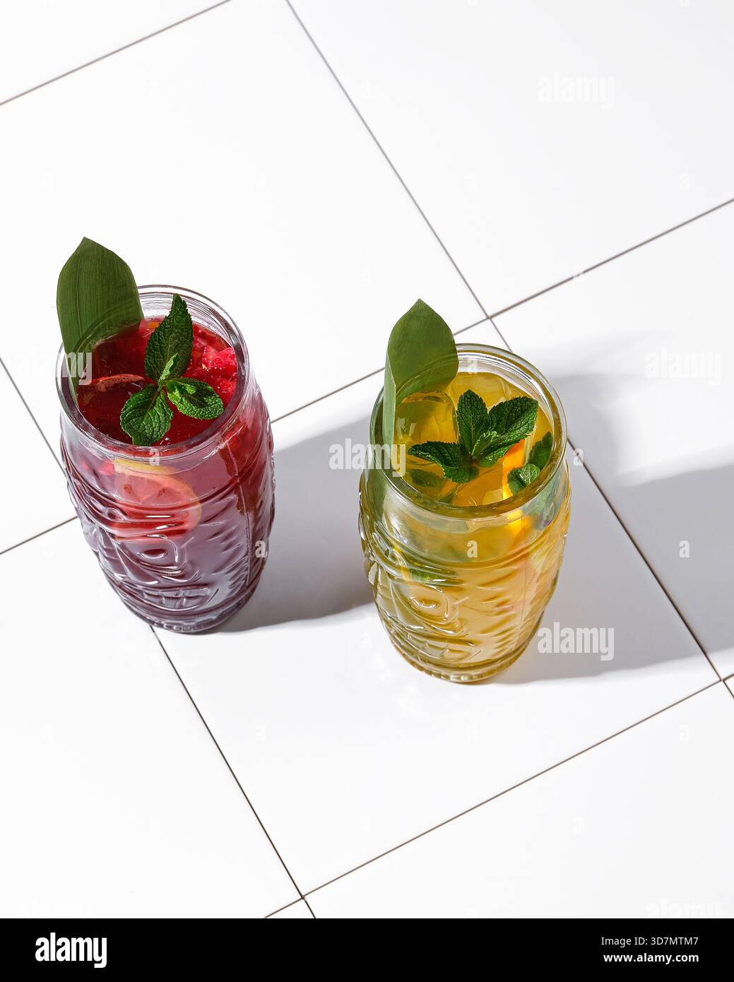 Zwei schattige Cocktails auf weißen Fliesen, rote und gelbe Gläser mit Minze- und großen Blattgarnituren, starke diagonale Schatten, sonnendurchflutete Gläser Stockfoto