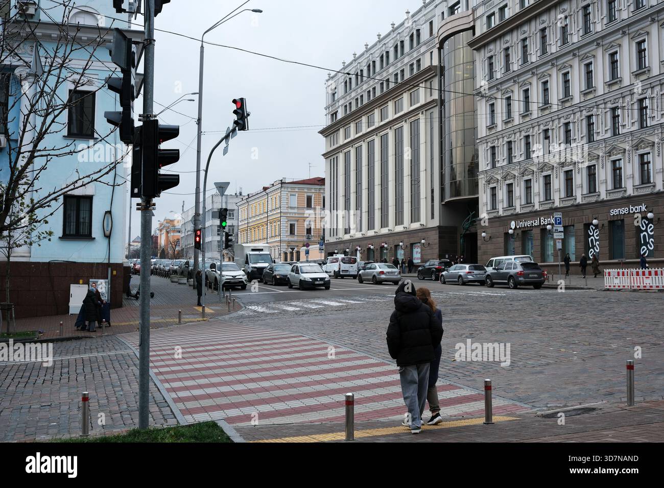 Zwei Leute laufen eine Straße entlang, während Autos an einer Ampel in der Ferne warten. Kiew, Ukraine, 26. November 2025. Stockfoto