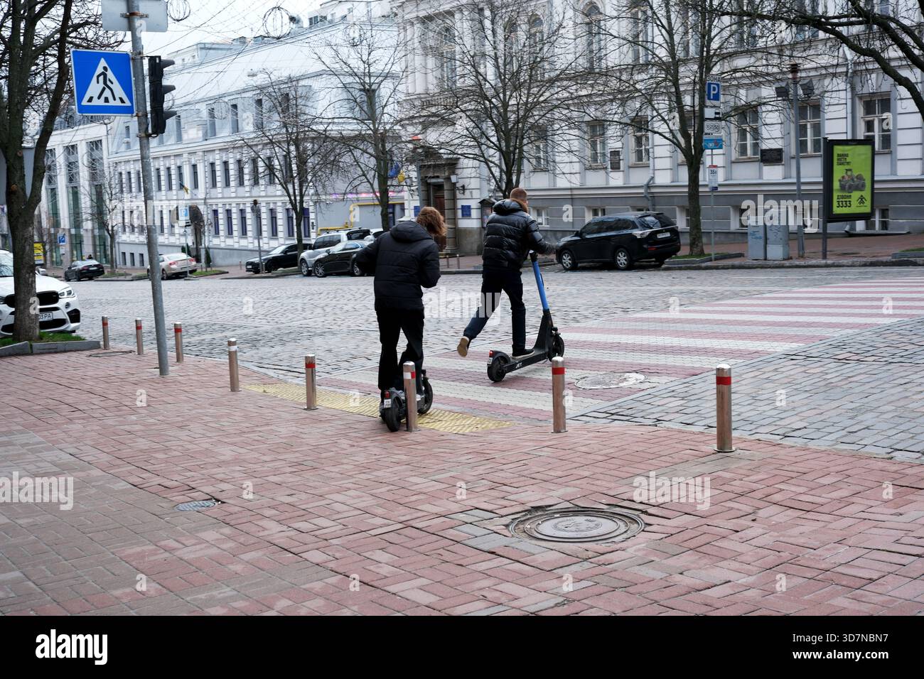 Zwei Personen fahren Elektroroller entlang einer ruhigen Straße im Morgenlicht. Kiew, Ukraine, 26. November 2025. Stockfoto