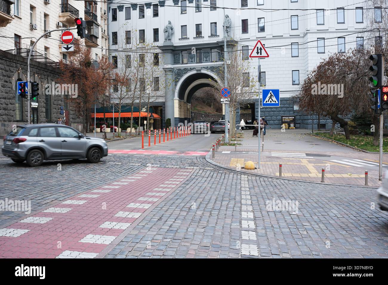 Autos fahren an einem bewölkten Tag durch eine kopfsteingepflasterte Kreuzung in der Nähe klassischer Gebäude. Kiew, Ukraine. November 2025. Stockfoto