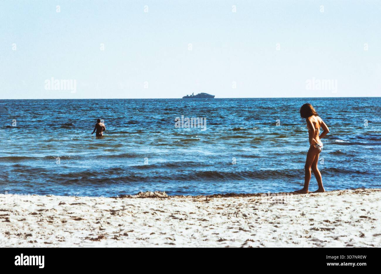 Rügen, Ddr. August 1987. Am Horizont ist ein Marinepatrouillenschiff der NVA sichtbar, das die DDR-Grenze auf dem Seeweg patrouilliert, während Menschen an Land in der Ostsee schwimmen Stockfoto