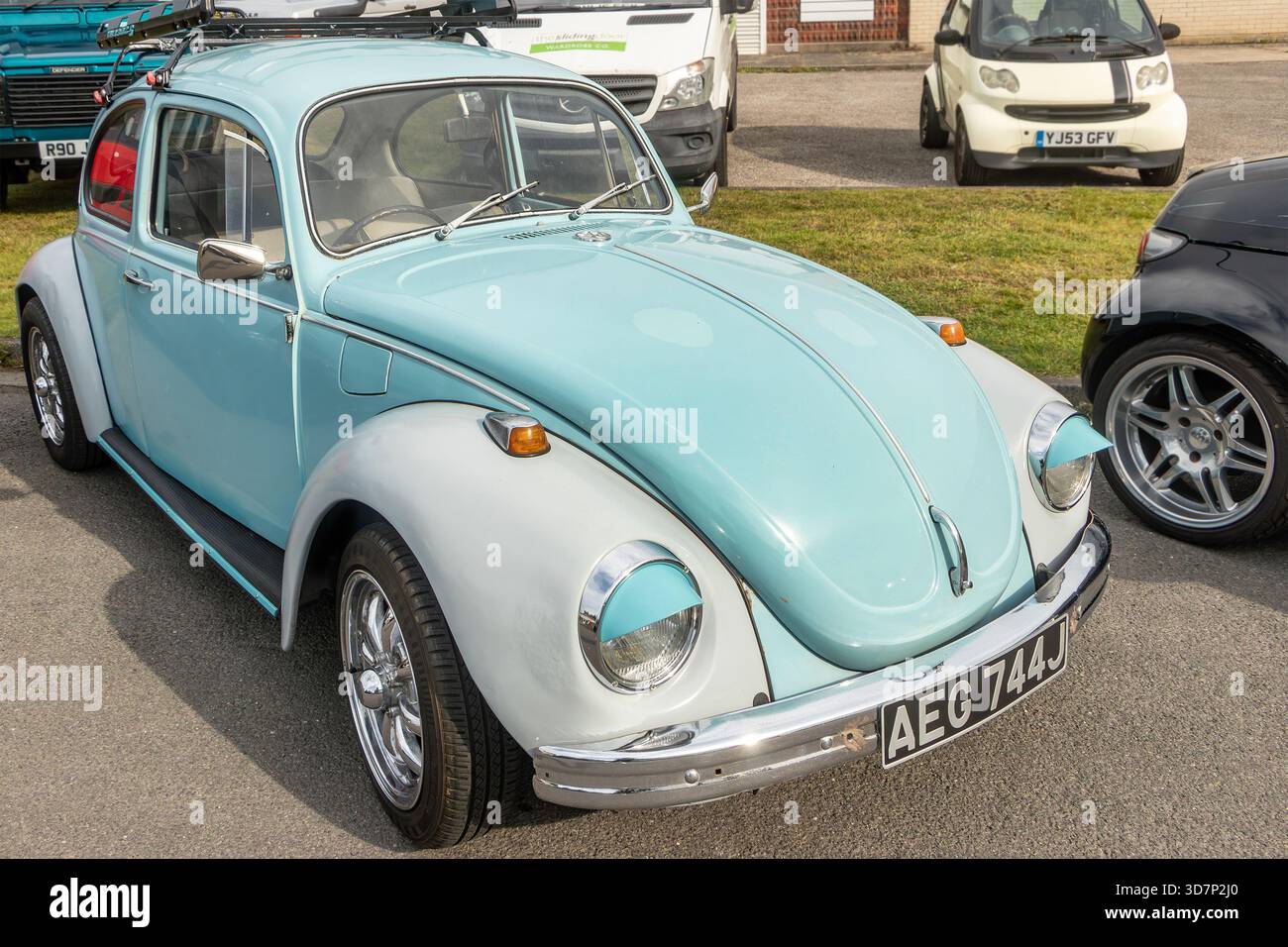 Der Volkswagen Beetle, der an einem sonnigen Tag bei einer Autoversammlungsveranstaltung neben anderen Fahrzeugen auf einem Bürgersteig geparkt wurde, war hellblau und weiß Stockfoto