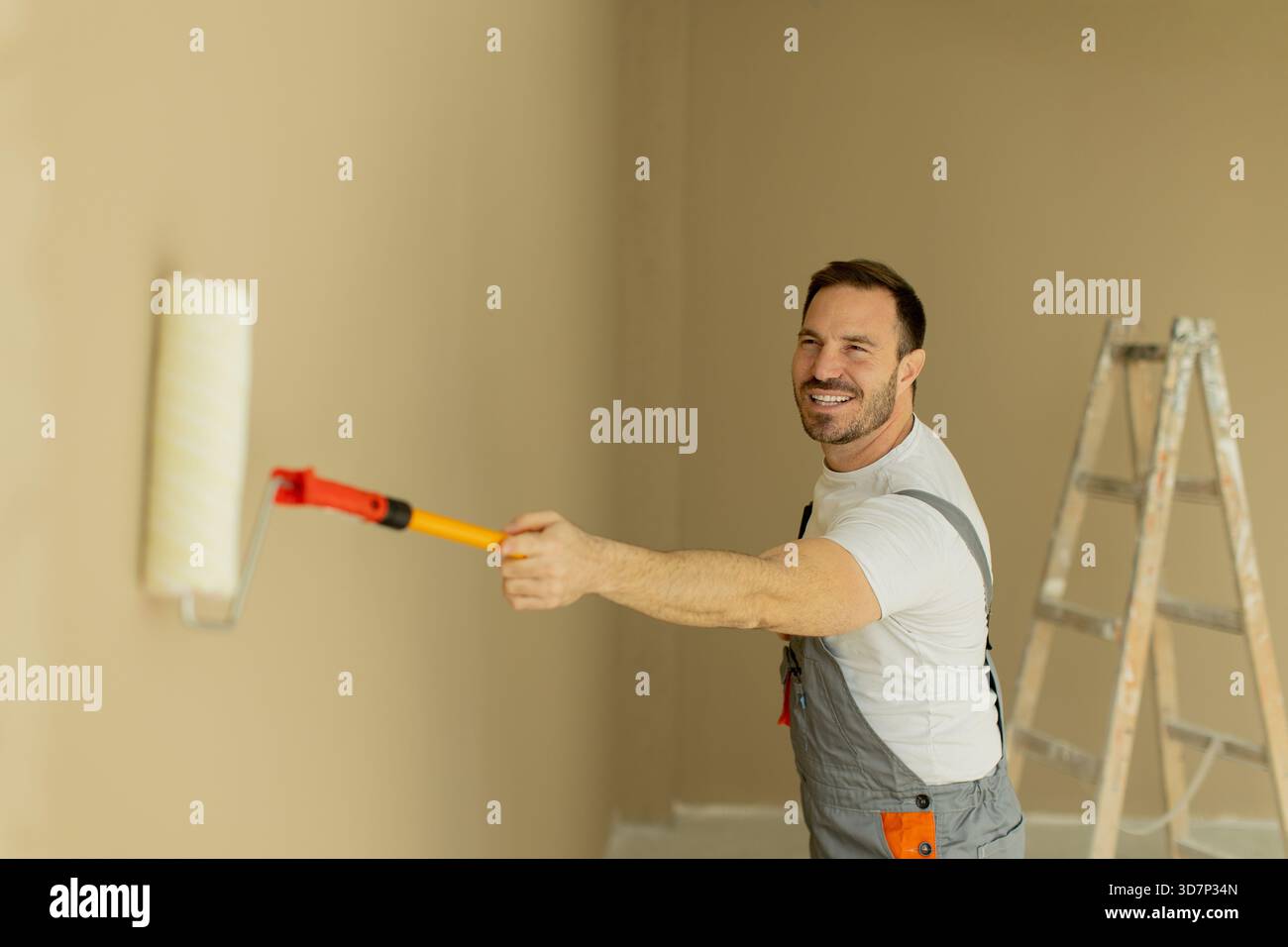 Ein Mann wälzt glücklich Farbe auf eine hellbeige Wand in einem Haus. Er trägt ein T-Shirt und einen Overall, mit einer Leiter in der Nähe und konzentriert sich auf die Aufgabe bei Stockfoto