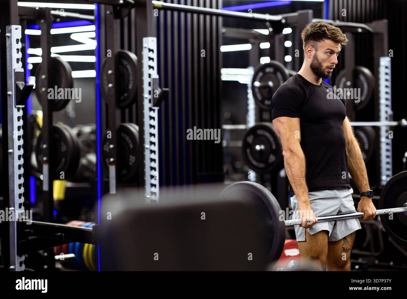 Ein männlicher Lifter führt einen Deadlift in einem gut ausgestatteten Fitnessstudio durch. Er konzentriert sich auf seine Form, während er eine schwere Langhantel hebt. Der Fitnessraum ist modern ausgestattet Stockfoto