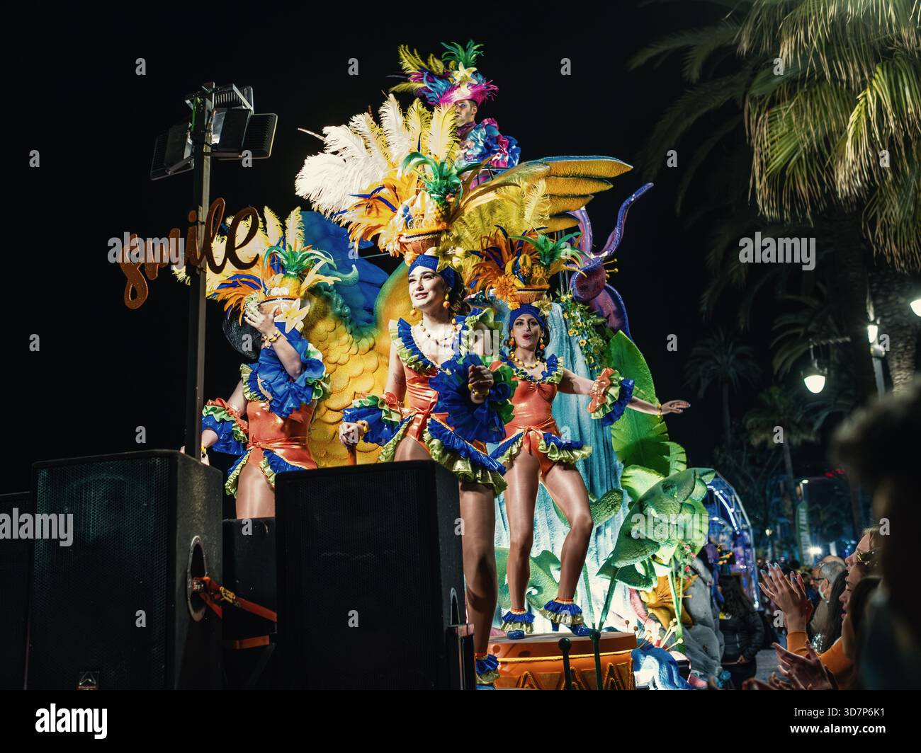Drei Erwachsene Frauen tragen während der Nachtparade des Karnevals 2022 leuchtende Federkostüme und Make-up-Tanz auf einem hell geschmückten Wagen in Sitges. Stockfoto
