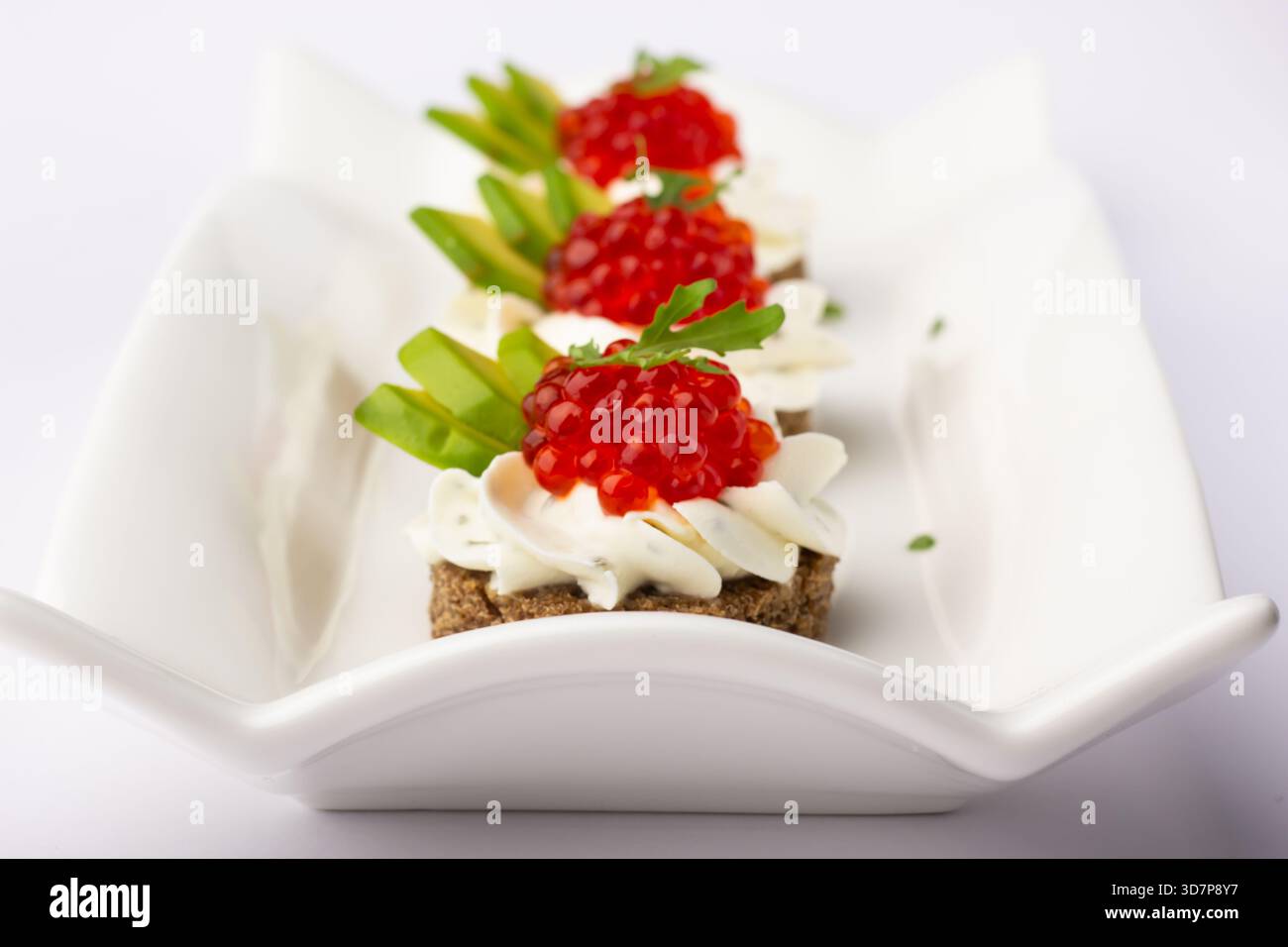 Drei köstliche Canapés mit Creme, farbenfrohem Kaviar und frischem Gemüse werden auf einem eleganten weißen Teller präsentiert. Diese elegante Vorspeise ist perfe Stockfoto
