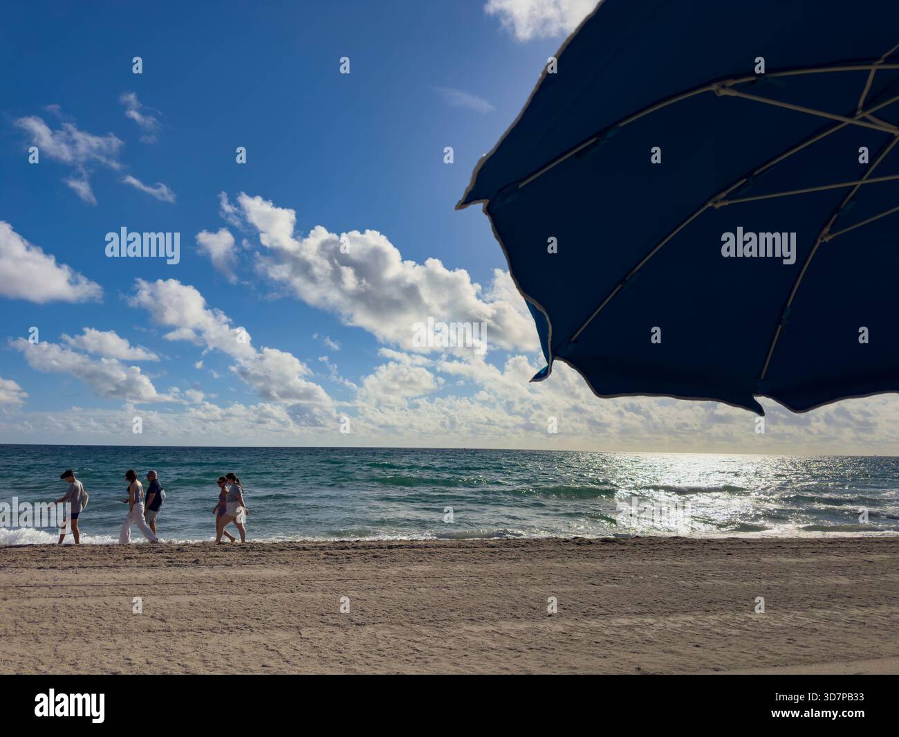 Strandbesucher spazieren an einem sonnigen Tag in Lauderdale-by-the-Sea unter einem hellen Morgenhimmel entlang der Küste, umrahmt vom Schatten eines großen Sonnenschirms - Smartphone-aufgenommenes Stockfoto