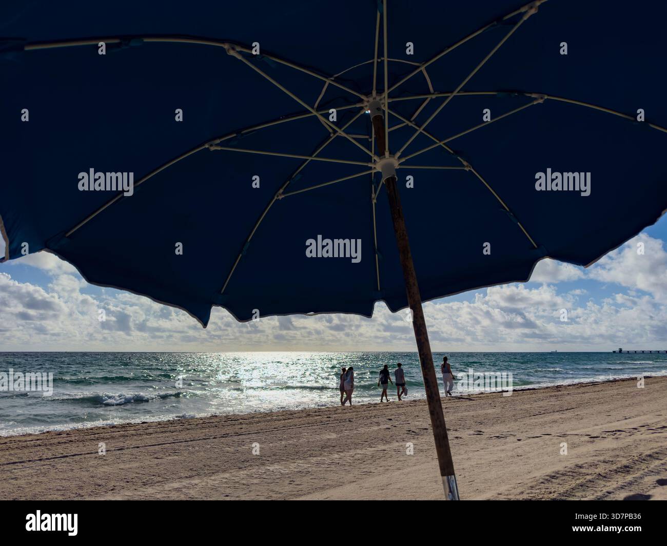 Strandbesucher spazieren an einem sonnigen Tag in Lauderdale-by-the-Sea unter einem hellen Morgenhimmel entlang der Küste, umrahmt vom Schatten eines großen Sonnenschirms - Smartphone-aufgenommenes Stockfoto