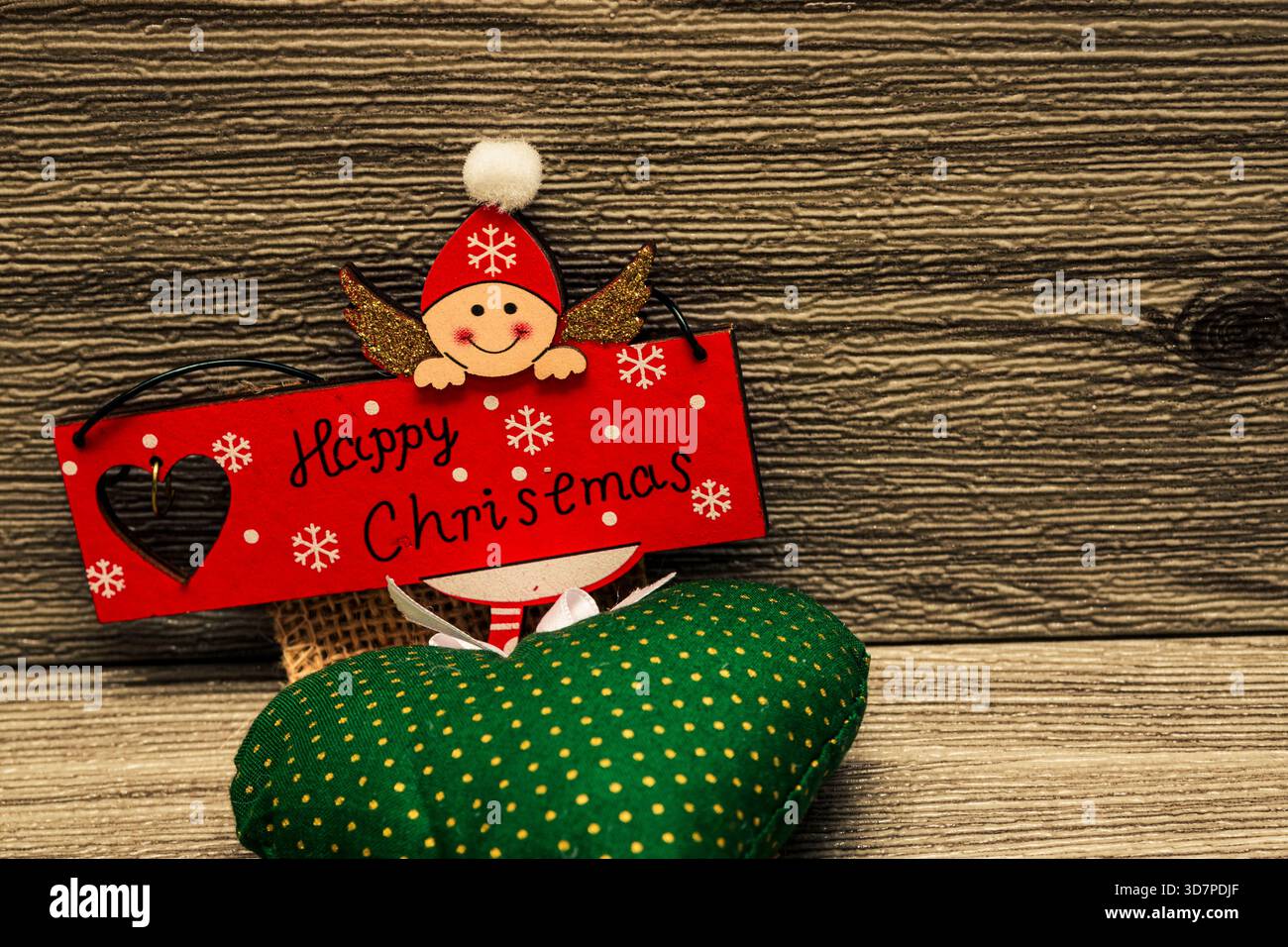 Niedlicher Holzengel mit rotem Weihnachtsschild über grünem Kissen auf rustikalem Holz, verspielte Weihnachtsdekoration. Stockfoto