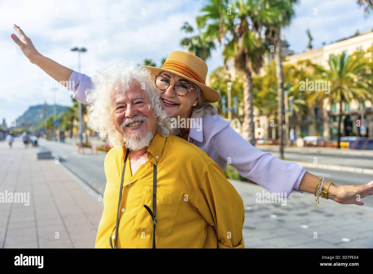 Seniorenpaar lacht, während der Ehemann seiner Frau einen Huckepack durch sonnendurchflutete Straßen der Stadt gibt, die von Palmen gesäumt sind, und genießen einen unbeschwerten Urlaub zusammen und Stockfoto