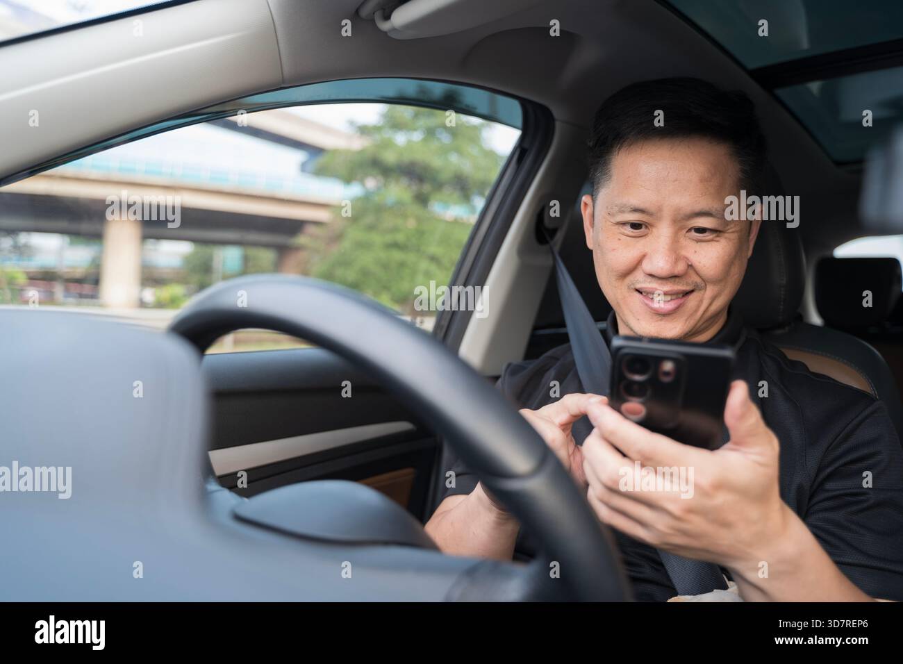 Ein Mann, der auf dem Fahrersitz sitzt, verwendet sein Smartphone, während das Fahrzeug angehalten wird, und hebt abgelenktes Fahrverhalten, den modernen Fahrzeuginnenraum und die hervor Stockfoto