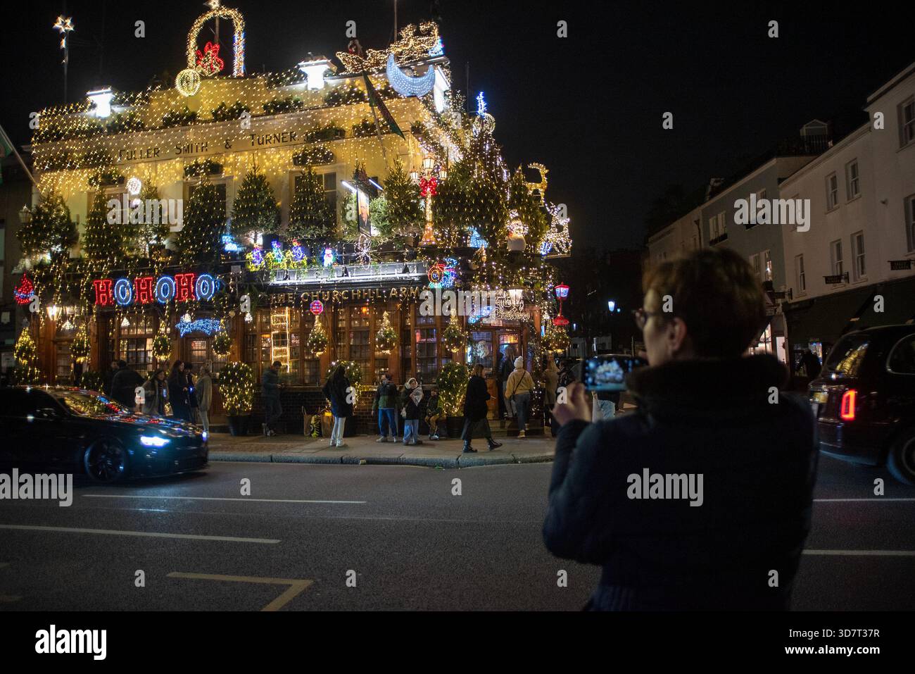 London, Großbritannien. November 2025. Ein Tourist macht ein Foto im Churchill Arms. Das Churchill Arms befindet sich in der Nähe der Notting Hill Gate Station. Es ist der weihnachtlichste Pub seit über 30 Jahren. In diesem Jahr wurden fast 100 Nadelbäume und 135000 Lichter verwendet, um den Pub draußen zu dekorieren. Quelle: SOPA Images Limited/Alamy Live News Stockfoto