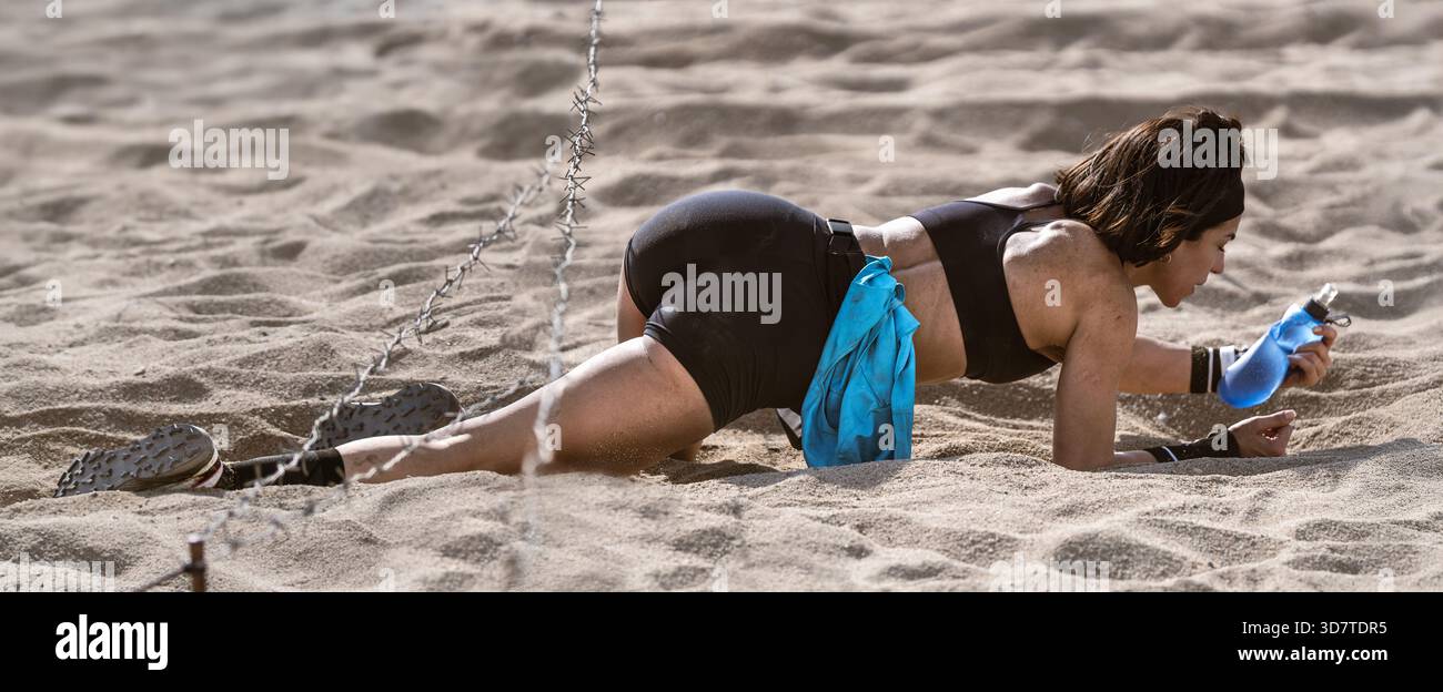 Weitwinkel-Nahaufnahme einer Athletin in eng anliegenden Shorts, die während eines Exters mit ihrer Wasserflasche unter Stacheldraht über den Sandstrand krabbelt Stockfoto