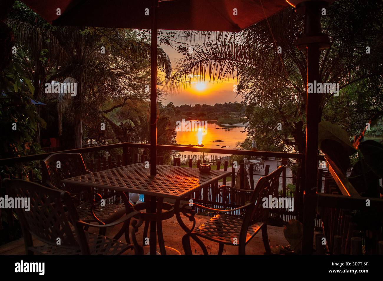 Ein leuchtend orangefarbener Sonnenuntergang, eingerahmt von Tisch, Stühlen und Vegetation auf dem Fluss Kavango - Verandah-Aufnahme. Stockfoto