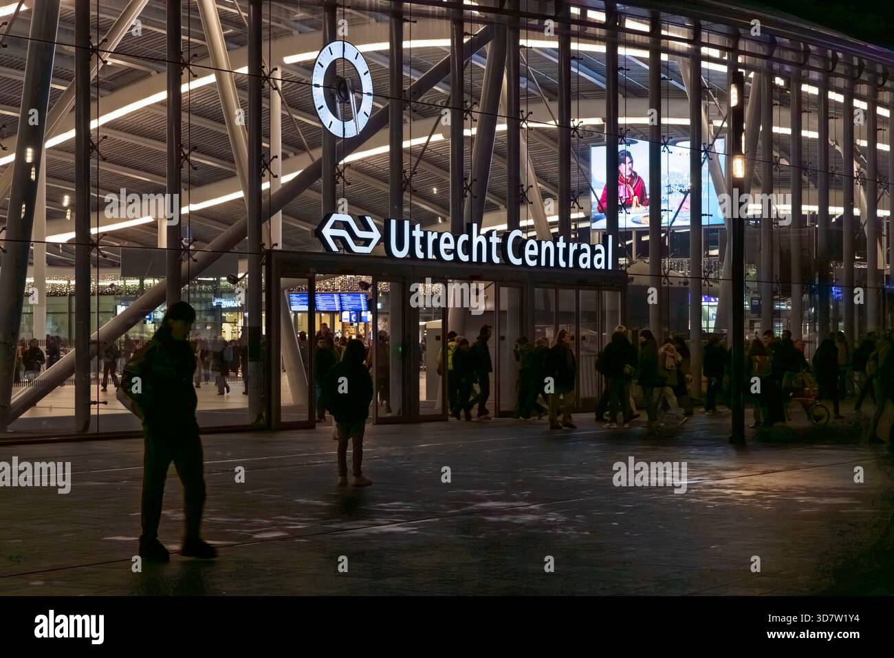 Die moderne Glasfassade und der Eingang zum Bahnhof Utrecht Centraal bei Nacht. Menschen, die an dem beleuchteten Schild des wichtigsten niederländischen Eisenbahnknotens vorbeilaufen. Utrec Stockfoto