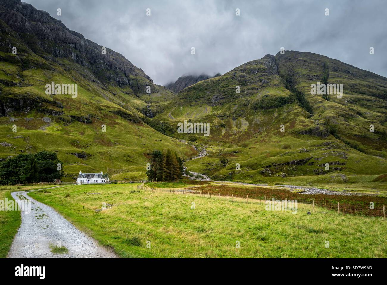 Ruhige Landschaft eines grünen Tals mit einem weißen Häuschen und majestätischen Bergen unter einem bewölkten Himmel. Glencoe, Schottland Stockfoto