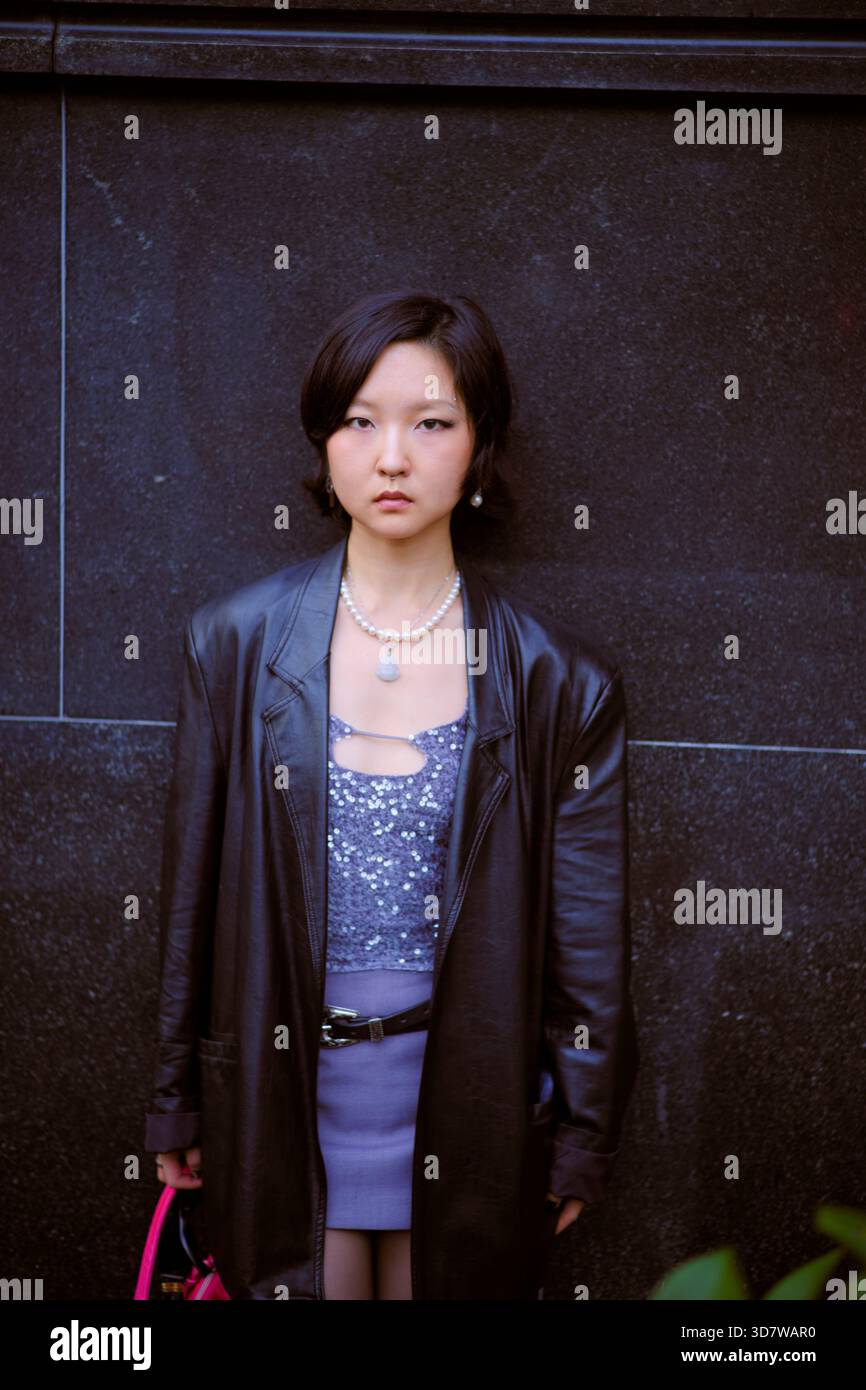 Eine junge Frau in stilvollem Outfit steht mit ernstem Ausdruck vor einer dunklen Steinmauer. Shanghai, China Stockfoto