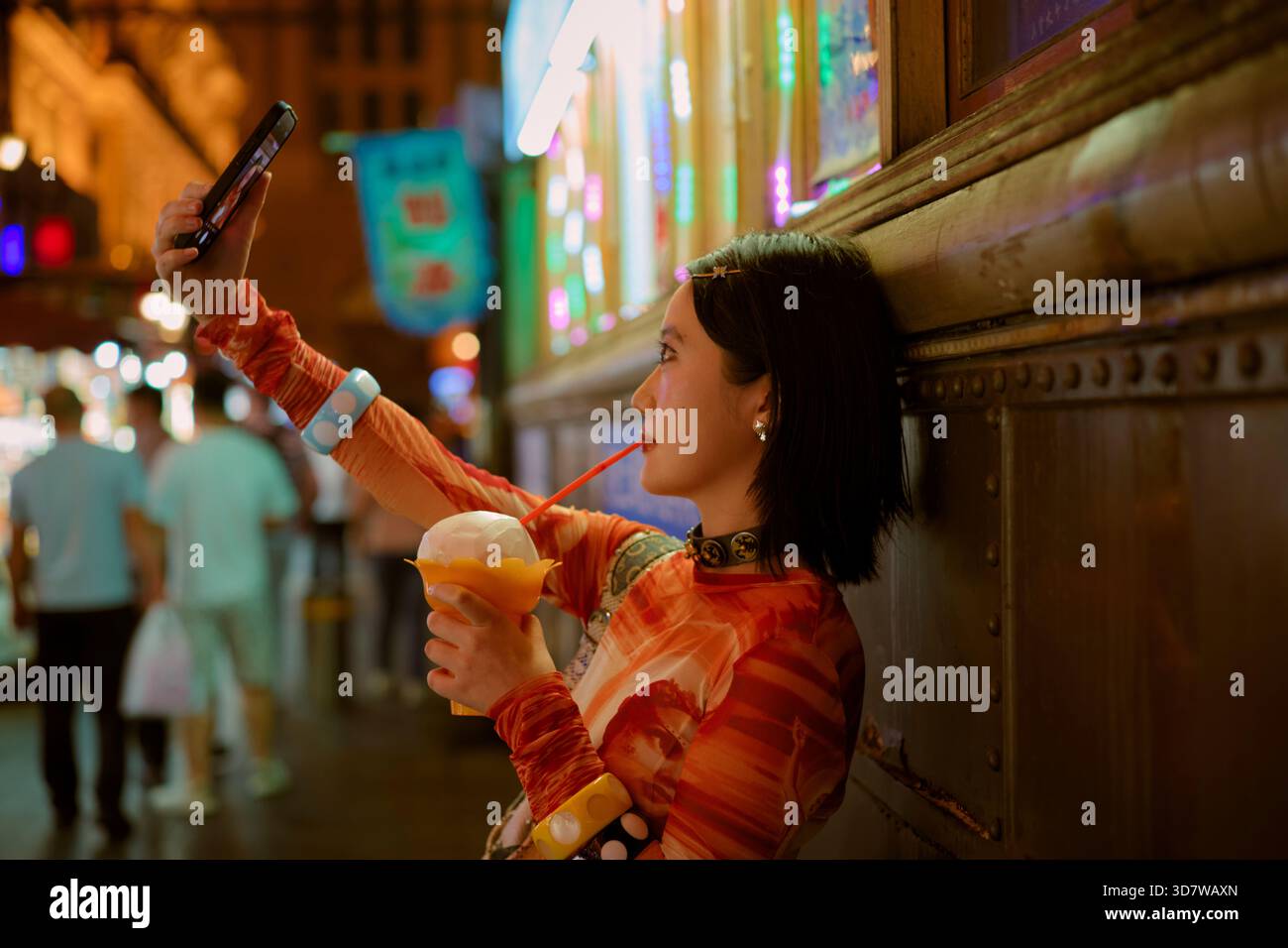Eine Frau, die nachts ein Selfie in einer lebhaften Stadtstraße macht und mit einem Strohhalm einen Drink hält. Shanghai, China Stockfoto