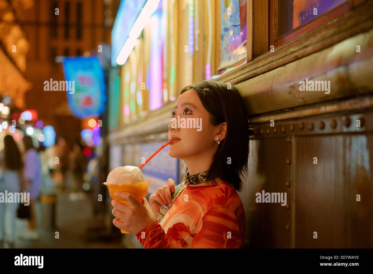 Frau in Orange genießt einen Drink mit einem Stroh in lebhafter, neonbeleuchteter urbaner Umgebung. Shanghai, China Stockfoto