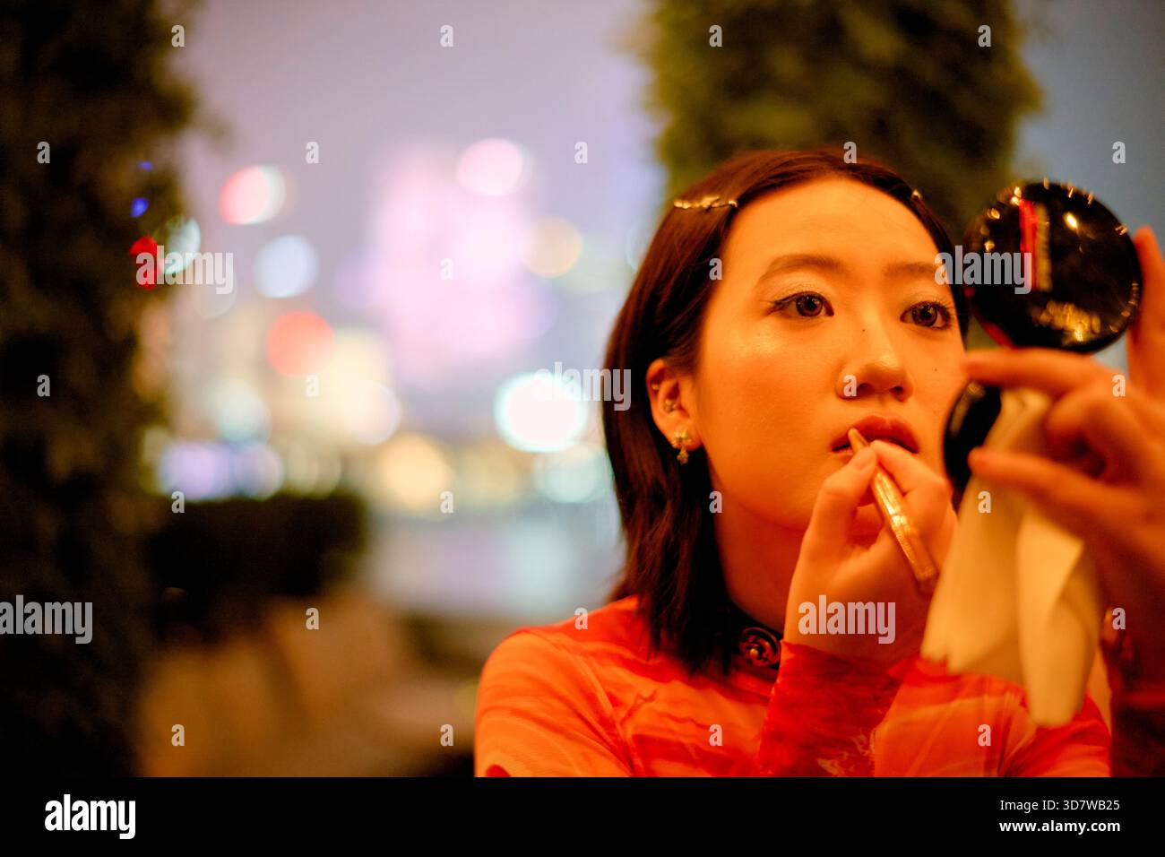 Junge Frau, die Lippenstift in einer Stadtlandschaft mit verschwommenen bunten Lichtern in der Nacht aufträgt. Shanghai, China Stockfoto