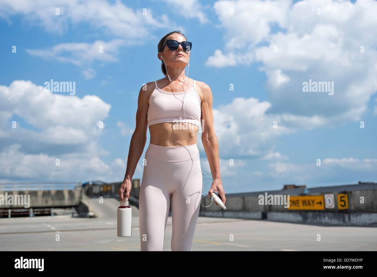 Sportliche Frau, die über einen Parkplatz läuft, Ohrstöpsel trägt und eine Trinkflasche trägt Stockfoto