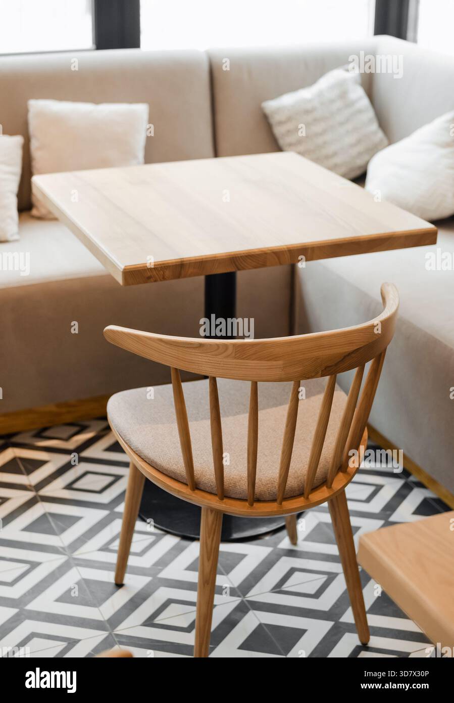 Gemütliches, modernes Café-Interieur. Heller Holztisch und passender Stuhl vor einem beigefarbenen Sitzsofa mit Kissen. Geometrisches Kachelmuster in Schwarz und weiß. Stockfoto
