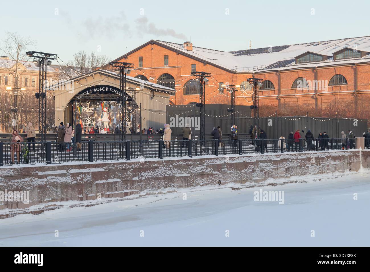 St-Petersburg, Russland - 7. Januar 2022: Eine lebhafte Winterszene entlang eines verschneiten Kanals bietet eine Eislaufbahn im Freien, eine festliche Bühne und Besucher in warmen Farben Stockfoto