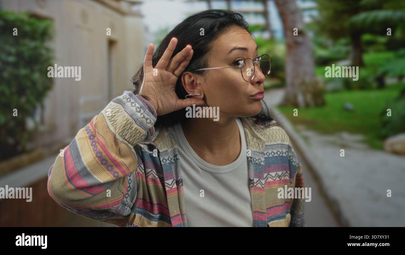 Frau, die Hand an Ohr schröpft, indem sie auf dem Parkweg baut, Brille und gemusterte Jacke trägt; Neugier hört aufmerksam zu. Stockfoto