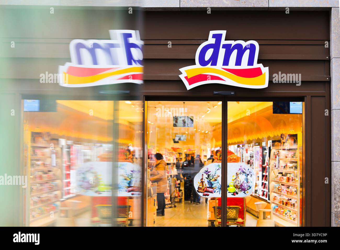 Bamberg, Deutschland 27. November 2025: Im Bild: Blick in den Eingangsbereich einer dm Drogerie in der Bamberger Innenstadt. Vor dem Geschäft kommt ein Fahrzeug oder Objekt vorbei und erzeugt Bewegungsunschärfen im Vordergrund, während sich die Kunden im Inneren bewegen Stockfoto