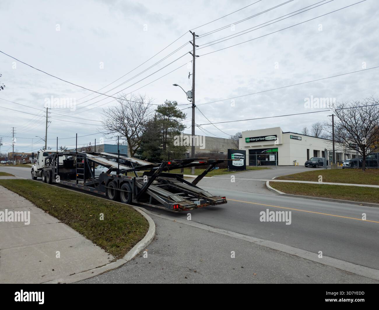 Leerer Autotransporter-Anhänger, der sich in der Nähe von Enterprise Rent-A-Car in der Industriezone Ajax unter bewölktem Winterhimmel abbiegt. - Smartphone-aufgenommenes Stockfoto