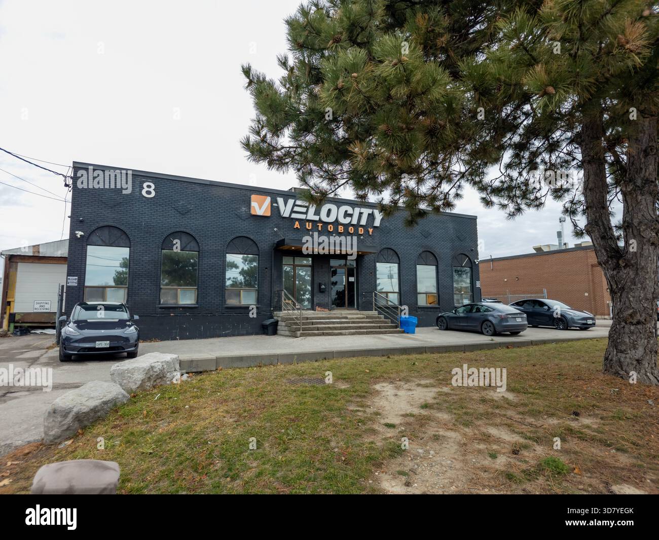 Velocity Autobody Geschäftsgebäude mit Bogenfenstern, geparkten Autos und auffälligen Schildern in urbaner Umgebung. - Smartphone-aufgenommenes Stockfoto