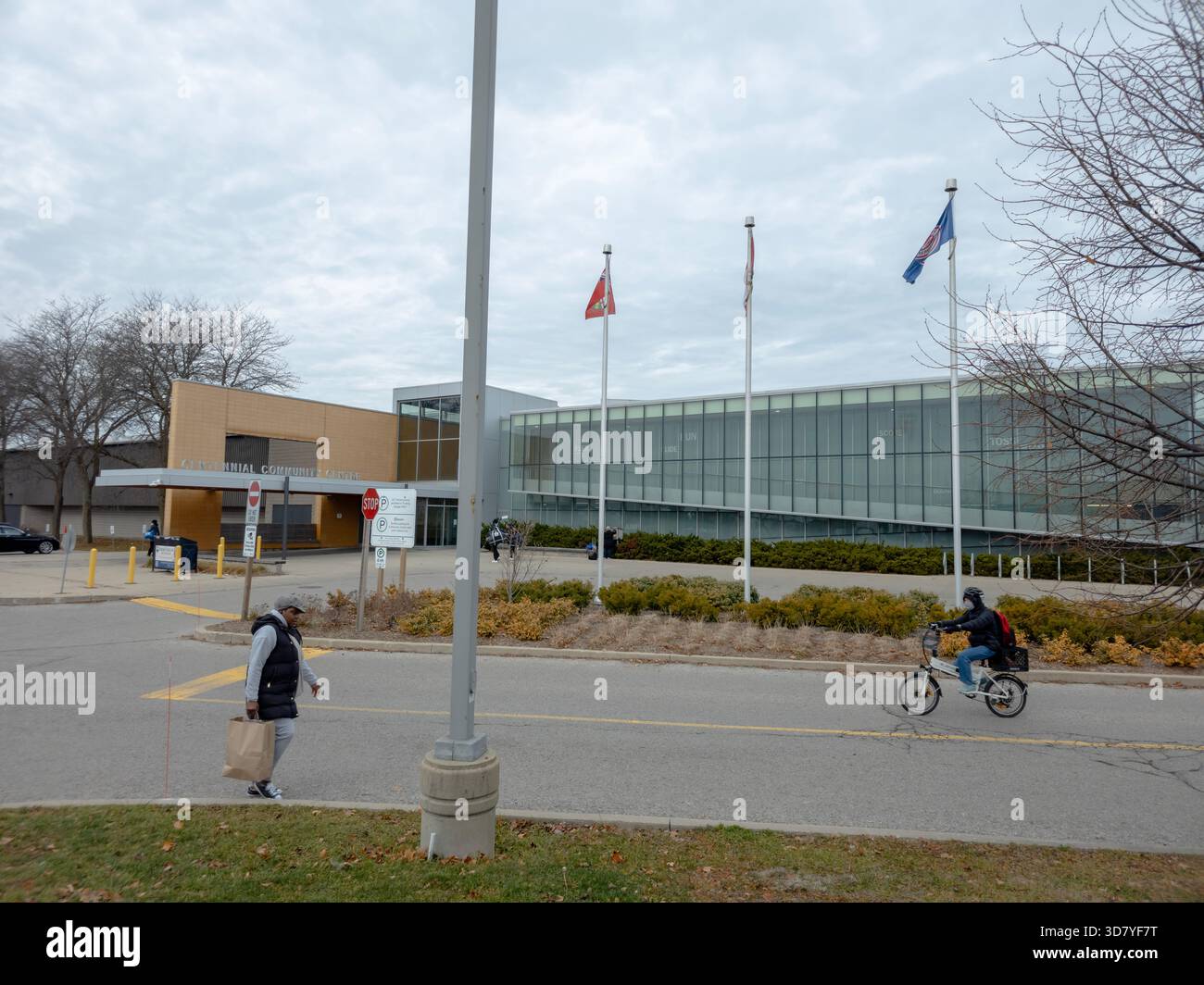 Modernes öffentliches Gebäude mit Flaggen, Glasfassade, Fußgängern, Radfahrern und landschaftlich gestaltetem Straßenrand unter bewölktem Himmel. gemeindezentrum. - Smartphone-aufgenommenes Stockfoto