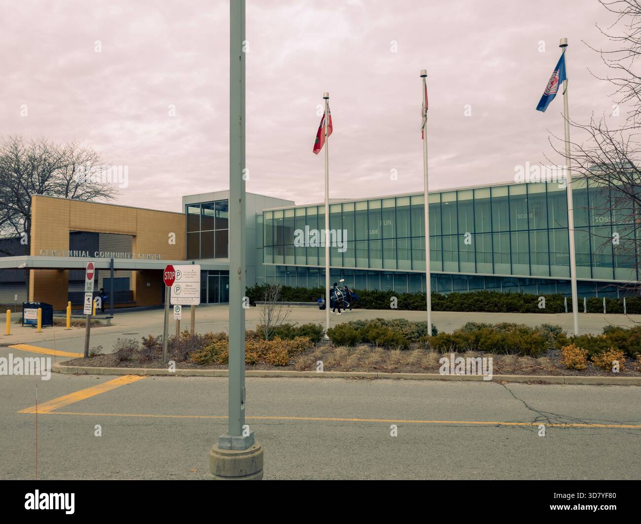 Modernes öffentliches Gebäude mit Flaggen, Glasfassade, Fußgängern, Radfahrern und landschaftlich gestaltetem Straßenrand unter bewölktem Himmel. gemeindezentrum. - Smartphone-aufgenommenes Stockfoto