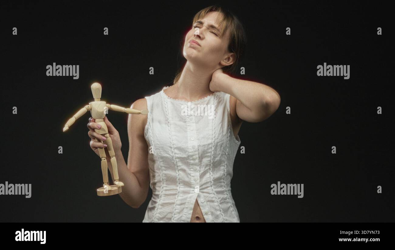 Junge Frau, die im schwarzen Atelier grimmt, während sie eine hölzerne Künstlerpuppe mit der Hand am Hals hält; Schmerzanfälligkeit. Stockfoto