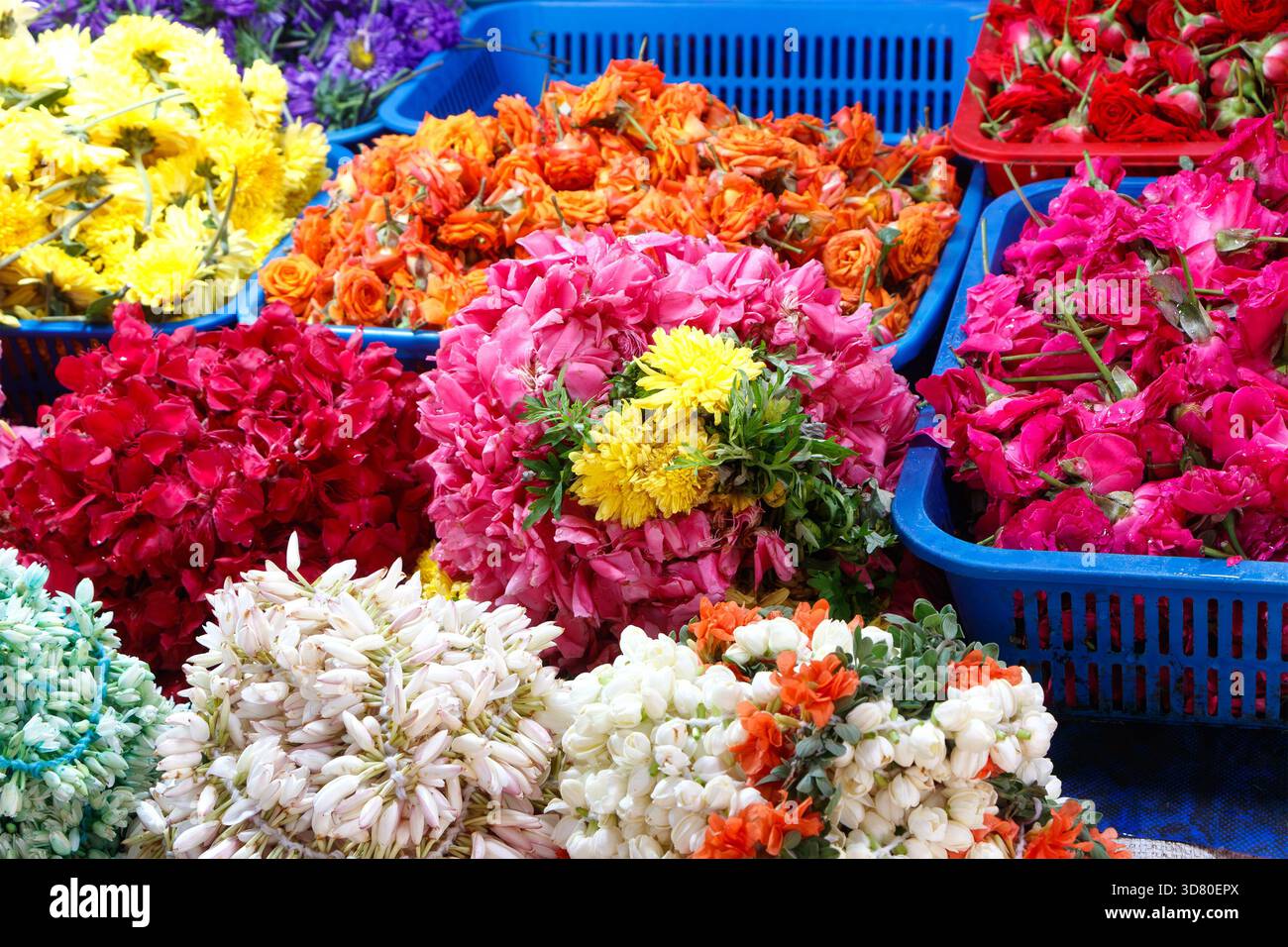 Farbenfrohe Auswahl an Blumengirlanden und losen Blüten in blauen Plastikkörben an einem lebhaften Marktstand im Freien Stockfoto