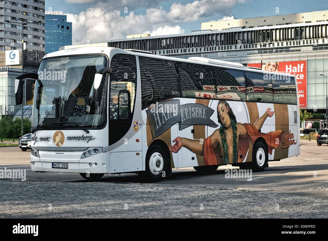 Weißer Bus mit einem großen Hare Krishna-Emblem auf der Seite, geparkt auf einem Stadtplatz, moderne Gebäude und Reflexionen sichtbar auf dem Glas. Stockfoto