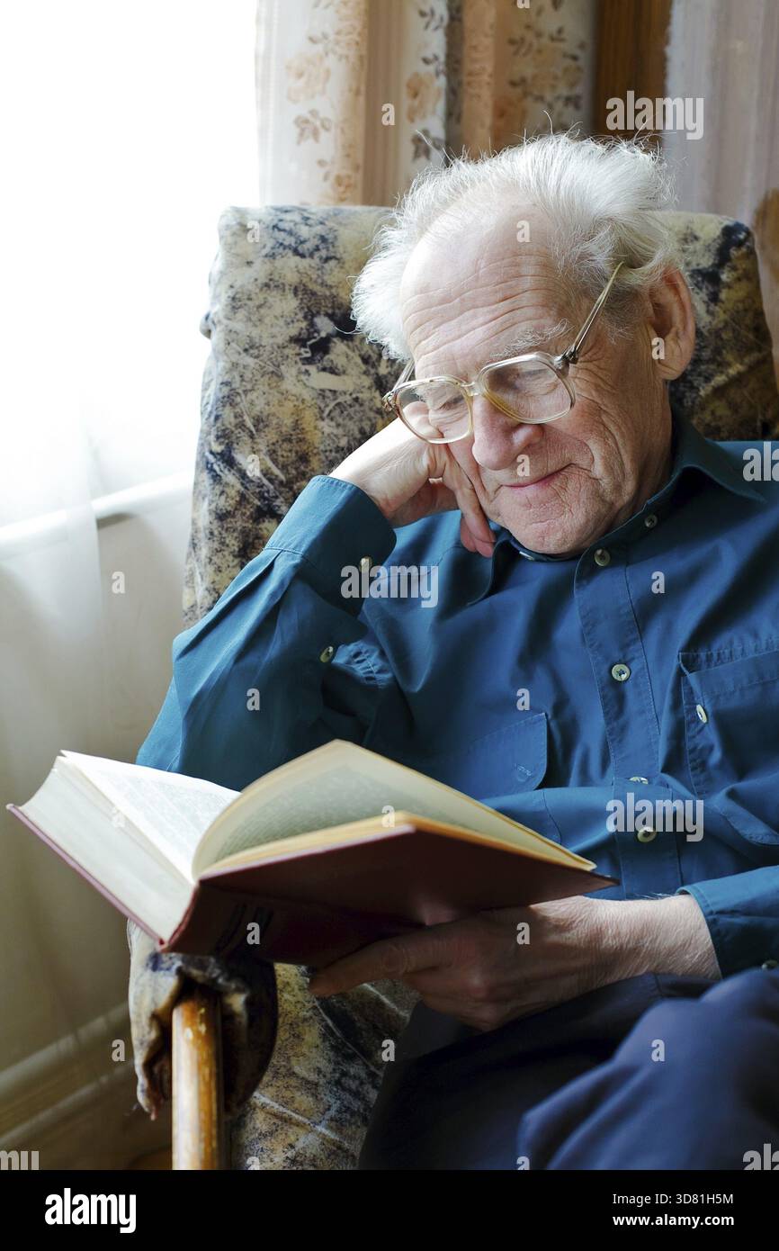 Ein lächelnder alter Mann in Brille liest ein lustiges Buch, hält den Kopf Stockfoto