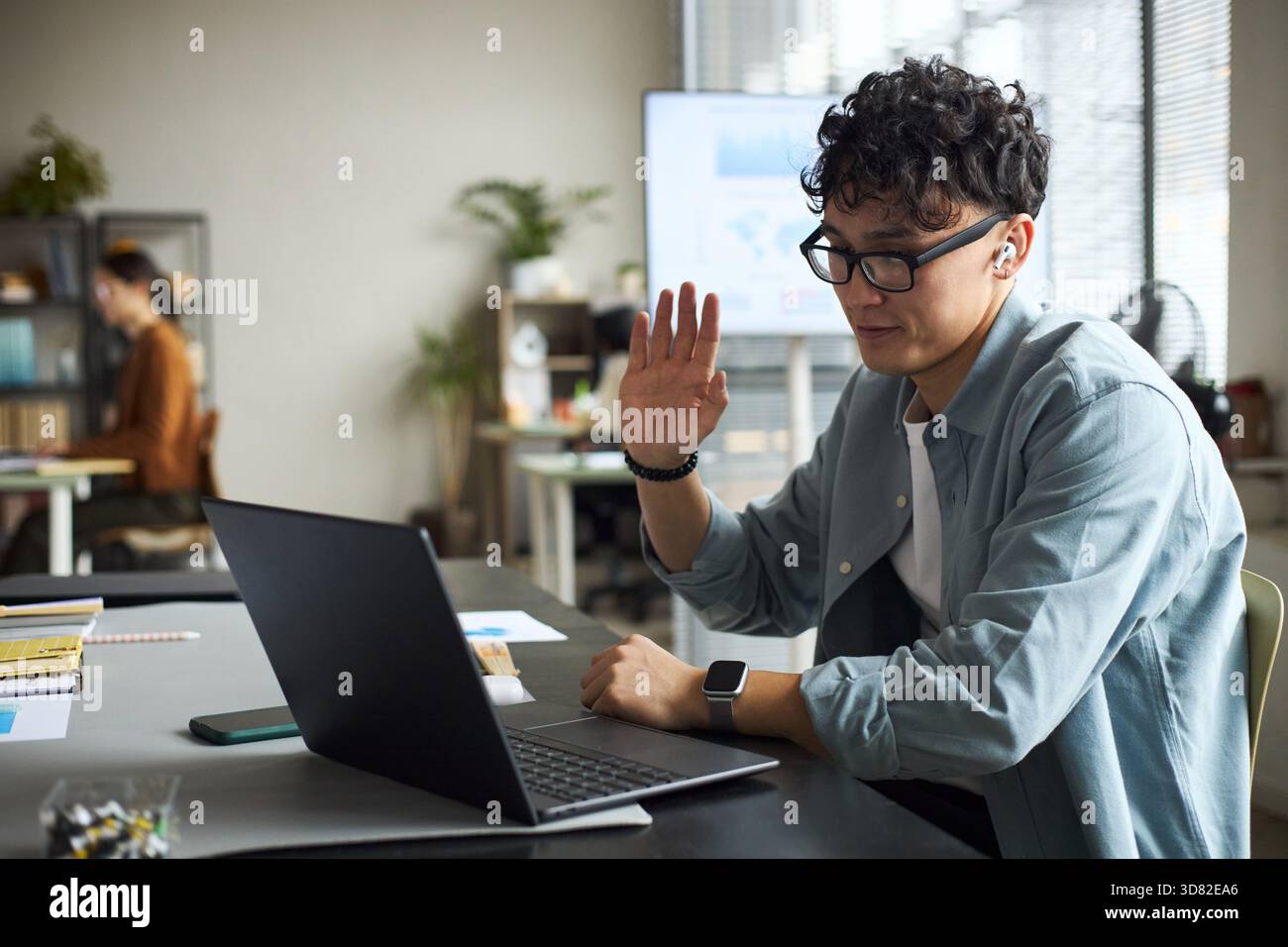 Junger asiatischer Mann mit Brille, der am Schreibtisch sitzt, mit Laptop die Hand hebt, während er an Videokonferenzen im modernen Büro teilnimmt, Geschäftsfrau, die im Hintergrund arbeitet Stockfoto