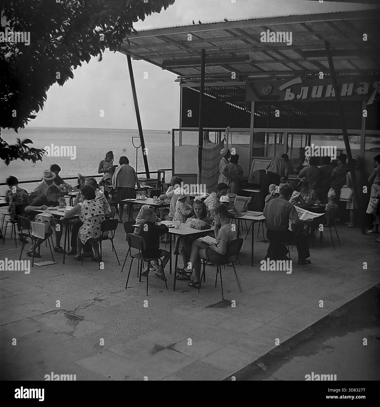 Krim, UdSSR - 1968: Eine geschäftige Szene auf einer Außenterrasse des „Blinnaya“ (Pfannkuchenhaus) mit Blick auf die Schwarzmeerküste. Urlauber essen an Metalltischen oder stehen in der Schlange am Serviceschalter unter einem Welldach. Eine Frau im gepunkteten Kleid isst zu Mittag, während ein Mann eine Zeitung im Vordergrund liest. Kinder sitzen in der Nähe und schauen sich ein Buch an. Dieses Schwarzweiß-Archivfoto zeigt die sowjetische Kultur der öffentlichen Gastronomie, die Freizeitatmosphäre und das tägliche Leben im berühmten Resort während der Sommerferien. Stockfoto