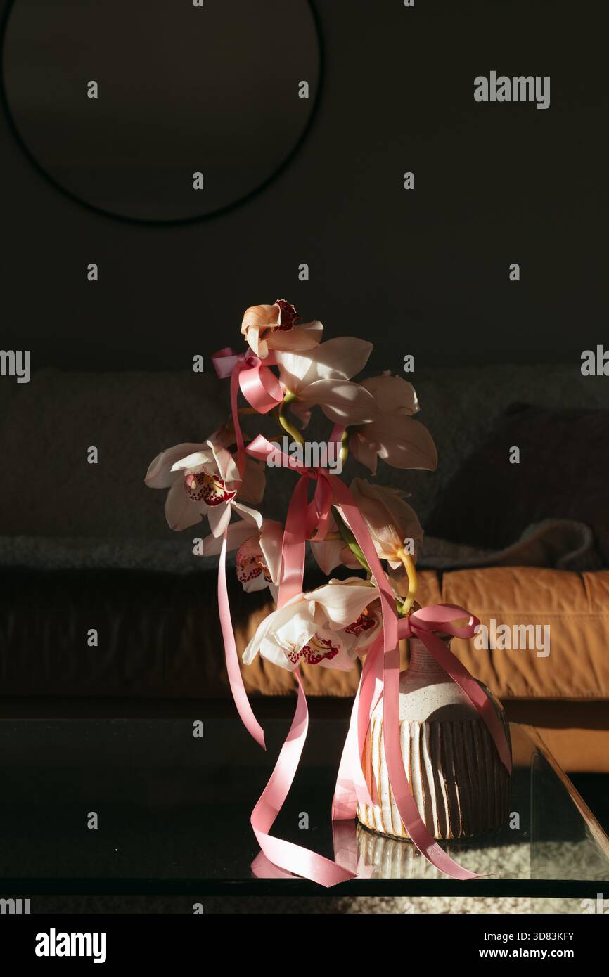 Weiße blühende Orchideen, gebunden mit rosa Schleife, stehend in einer strukturierten Vase auf einem Glastisch. Sonnenlicht erzeugt stimmungsvolle Schatten Stockfoto
