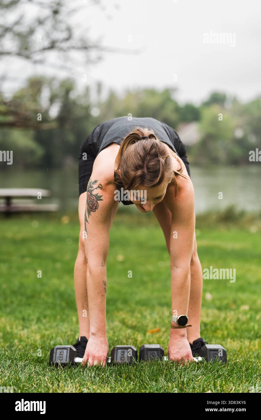 Frau, die sich beugt, um Kurzhanteln auf Gras in der Nähe eines Sees zu schnappen Stockfoto