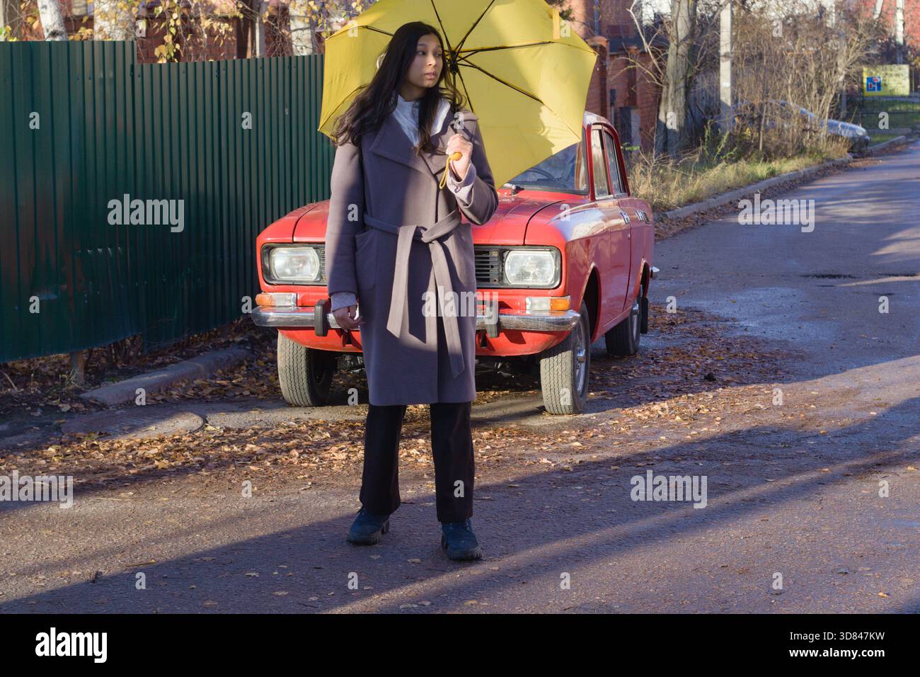 Eine junge Indianerin schlendert an einem roten Oldtimer vorbei, hält einen gelben Regenschirm und hält eine Hand in ihrer Jackentasche, Vorderansicht Stockfoto