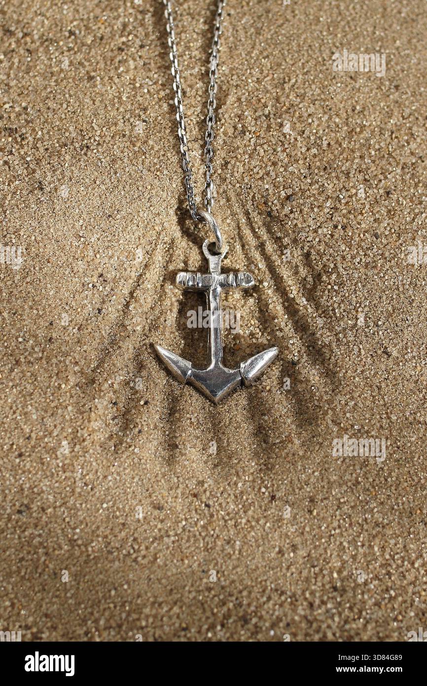 Anhänger mit silbernem Anker-Halskette, der auf Strandsand glänzt, symbolisiert Hoffnung und maritimes Abenteuer. Stockfoto