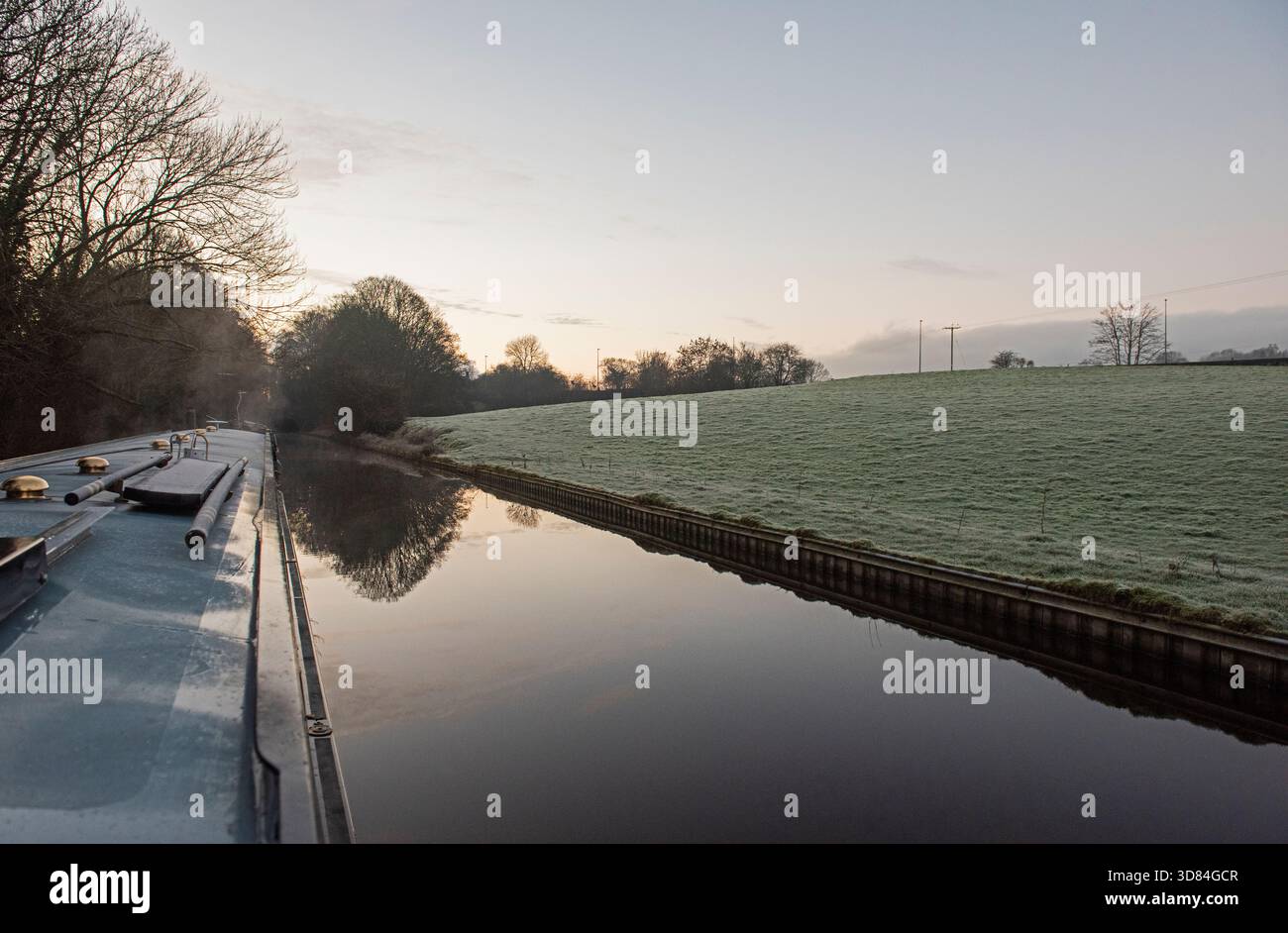 Blick aus dem Schmalboot auf die englische ländliche Landschaft auf einem britischen Wasserstraßenkanal mit Bäumen und Wanderweg im Winter am frostigen Morgen mit Sonnenaufgang Stockfoto