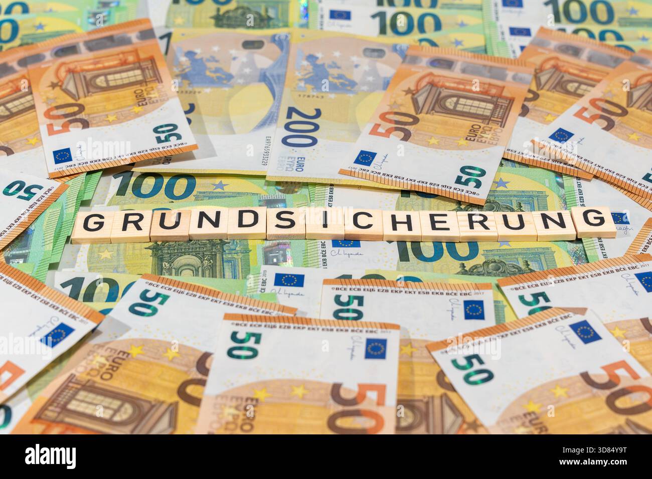 Grundsicherung erscheint in Holzfliesen auf gemischten Euro-Banknoten. Symbolisches Stillleben über Sozialhilfe, Lebenshaltungskosten und öffentliche Hilfe. Stockfoto