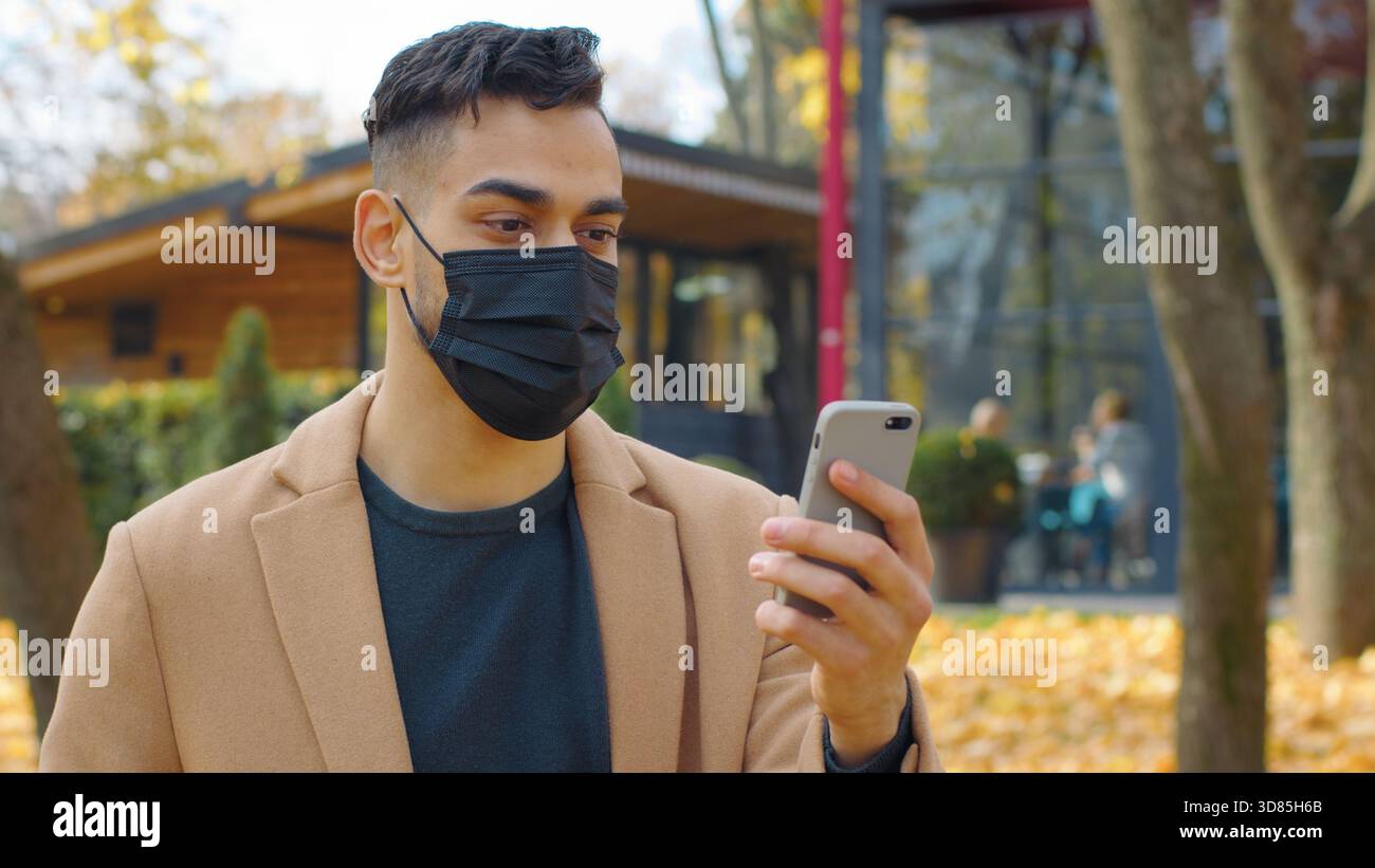 Hispanic Mann im Gesicht medizinische Maske in Handy mit App Smartphone im Park Herbst Herbst Saison Arabian Mann chatten Telefon sozial Stockfoto