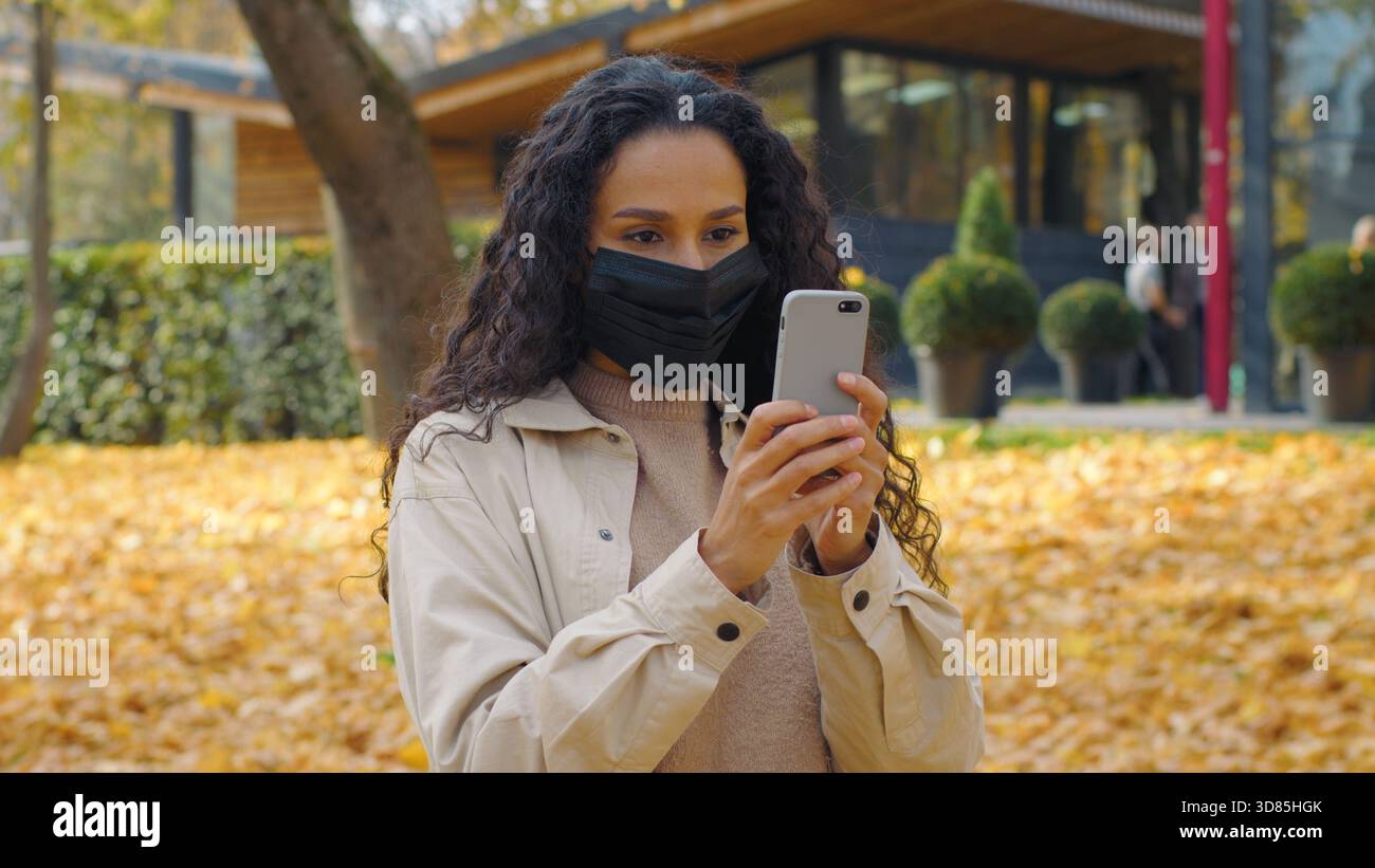 Arabian Frau Hispanic Mädchen im Gesicht medizinische Maske mit Handy im Herbst Park Dame Scrollen Smartphone App chatten online Internet Medien Stockfoto
